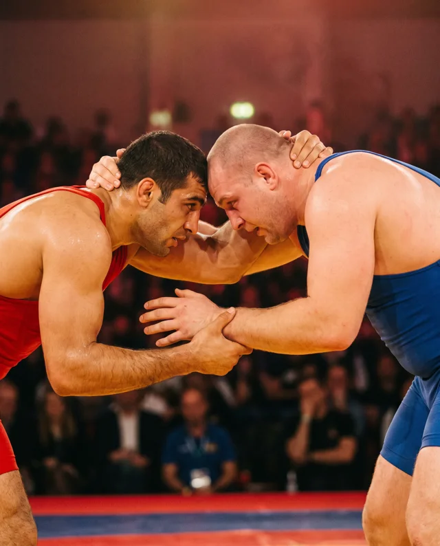 Two wrestlers in red and blue singlets locked head to head in a collar tie